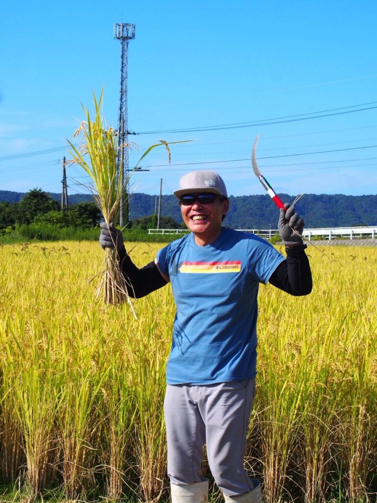 実った稲と鎌を手に笑顔を見せるスタッフ